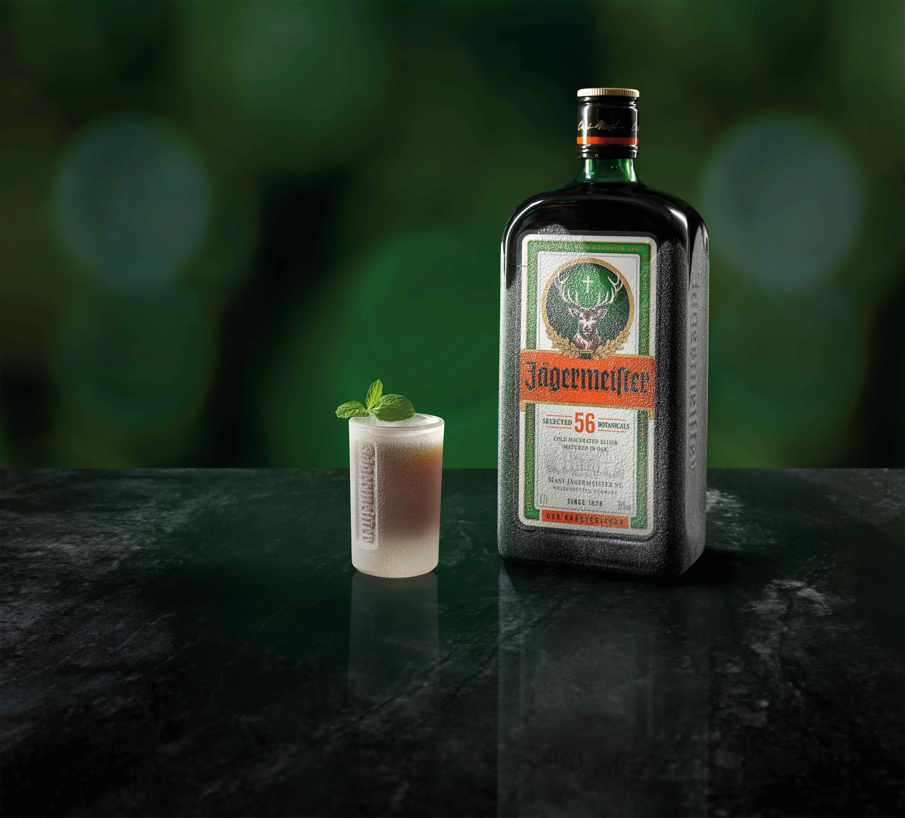 A ice cold shot glas of Jägermeister topped with min standing in front of a Jägermeister bottle